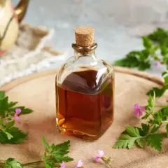 Szklana butelka z bursztynową nalewką z geranium, otoczona świeżymi gałązkami i kwiatami tej rośliny, na drewnianej desce.