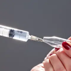Dłonie z ciemnoczerwonym lakierem przygotowują zastrzyki na opalanie, pobierając płyn z ampułki do strzykawki.