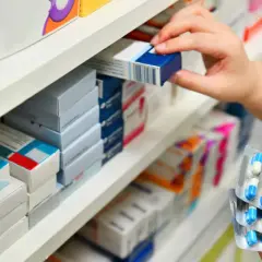 Ręka sięga po lek na aptecznej półce. W dłoni trzymane są blistry z niebiesko-białymi kapsułkami, symbolizujące moment, kiedy antybiotyk zaczyna działać.