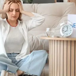 Kobieta z bólem głowy podczas menopauzy szuka leków na menopauzę po których się nie tyje. Na stoliku budzik i tabletki.