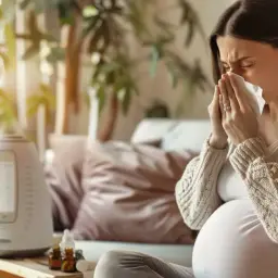 Ciężarna kobieta z chusteczką przy nosie, szukająca odpowiedzi na pytanie: co na bolące gardło w ciąży. Obok nawilżacz powietrza i buteleczki z lekami.