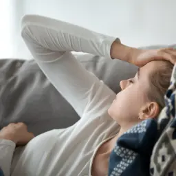 Kobieta leży na kanapie, trzymając się za czoło. Może to być jeden z wczesnych objawów ciąży, takich jak zmęczenie lub ból głowy.