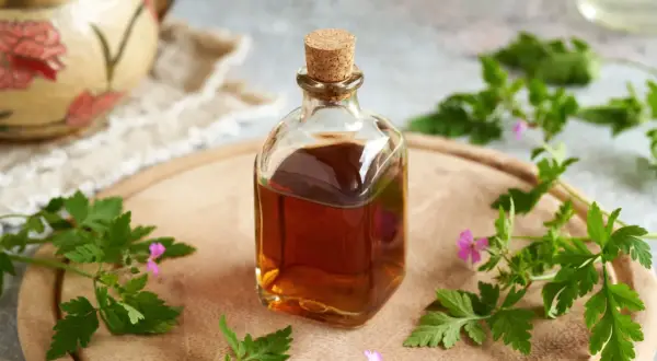 Szklana butelka z bursztynową nalewką z geranium, otoczona świeżymi gałązkami i kwiatami tej rośliny, na drewnianej desce.