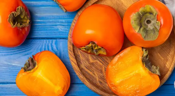 Pomarańczowe owoce kaki na niebieskim tle. Uważaj na potencjalne skutki uboczne spożycia, zwłaszcza na pusty żołądek.
